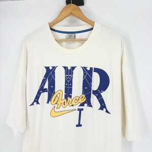 May include: Cream-colored t-shirt with a blue and yellow graphic. The design features the word "AIR" in blue, with a grid pattern, and "Force" in yellow, along with a yellow Nike swoosh. The shirt is hanging on a wooden hanger.