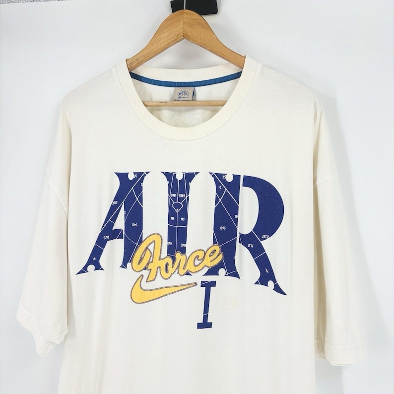 May include: Cream-colored t-shirt with a blue and yellow graphic. The design features the word "AIR" in blue, with a grid pattern, and "Force" in yellow, along with a yellow Nike swoosh. The shirt is hanging on a wooden hanger.