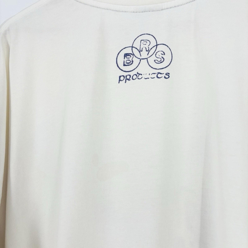 May include: Cream-colored long-sleeve shirt with a crew neck. The shirt features a navy blue graphic on the upper chest, consisting of three overlapping circles with the letters "B", "R", and "S" inside, and the word "products" below.