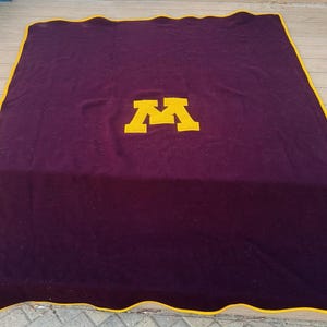 May include: A large, rectangular maroon blanket with a gold border. The blanket features a large gold letter "M" in the center. The blanket is spread out on a wooden surface.