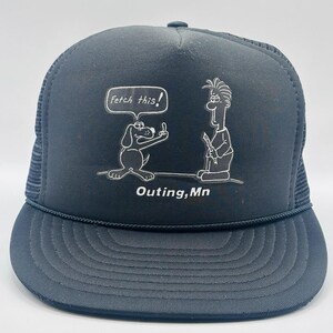 Puede incluir: Gorra de camionero azul marino con un dibujo blanco de un perro haciendo una peineta y el texto "Fetch this!". También hay una figura de dibujos animados. El texto "Outing, Mn" está debajo.