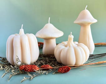 May include: Four ivory-colored candles in the shape of pumpkins and mushrooms. The candles are arranged on a bed of dried foliage and set against a teal background. The pumpkin candles have ribbed sides, and the mushroom candles have a rounded cap.