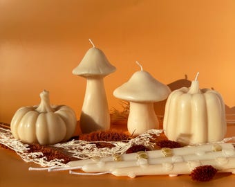 May include: A collection of ivory-colored candles in the shapes of pumpkins and mushrooms. The candles are arranged on a white lace doily with dried floral accents against an orange background. Several long, thin candles are also present.