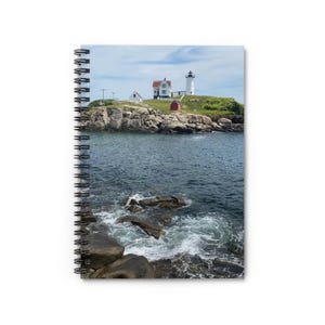 May include: Spiral-bound notebook with a coastal scene. The image depicts a lighthouse on a rocky island, with a red building and a white lighthouse tower. The sea surrounds the island, with waves breaking against the rocks. The sky is blue with some clouds.