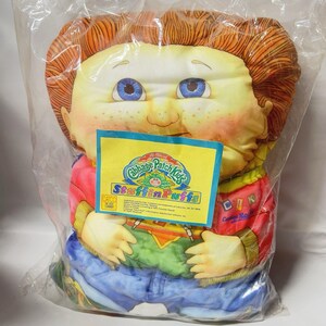 May include: A Cabbage Patch Kids StuffinPuffs plush toy featuring a character with orange hair, blue eyes, and freckles. The character wears a red jacket with blue pants and a green shirt. A yellow product tag is attached to the front of the plastic bag.