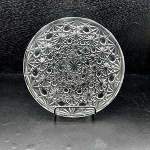 Puede incluir: Plato de vidrio transparente con un detallado patrón de estrella. El plato redondo se encuentra sobre un soporte de metal, mostrando un diseño vintage. El intrincado diseño crea una apariencia texturizada, perfecto para servir o exhibir.