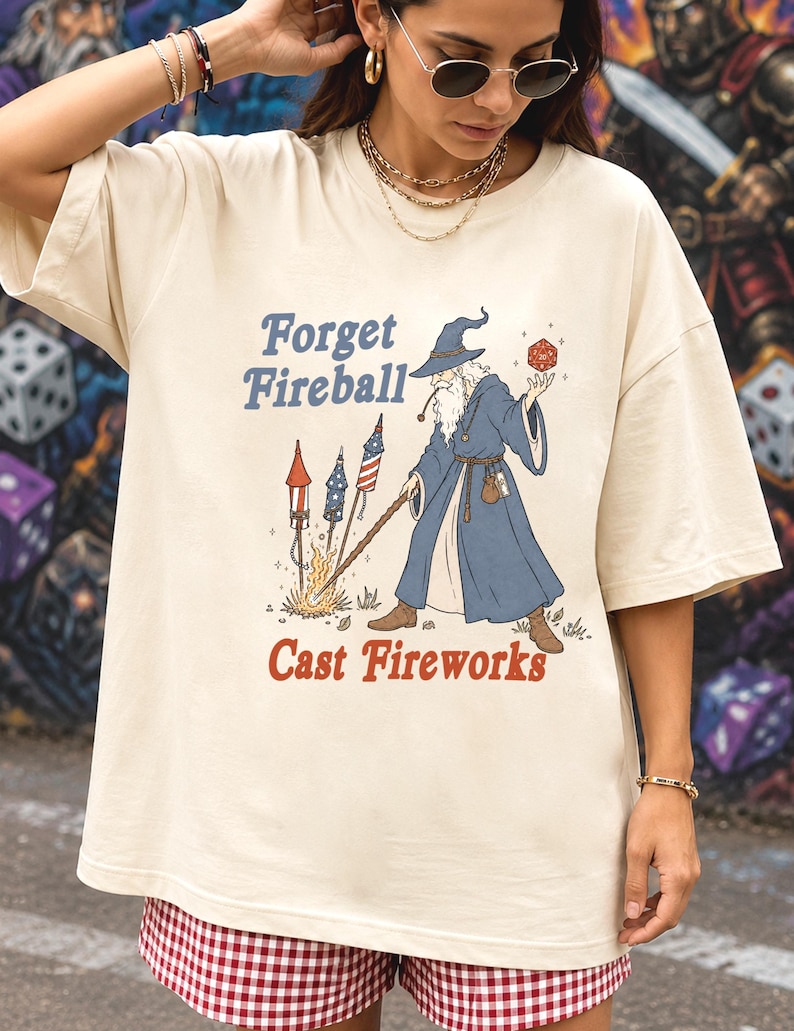 Op de afbeelding: Cr&egrave;mekleurig T-shirt met een afbeelding van een tovenaar die vuurwerk afsteekt. De tekst luidt "Forget Fireball Cast Fireworks" in blauw en rood. De tovenaar draagt een blauwe mantel en hoed en houdt een rode dobbelsteen vast.