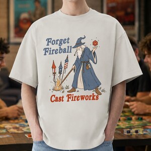 Op de afbeelding: Lichtbeige t-shirt met de tekst "Forget Fireball Cast Fireworks". De afbeelding toont een tovenaar in een blauwe mantel met een rode dobbelsteen en een staf, met vuurwerk. Een casual, unisex stijl.