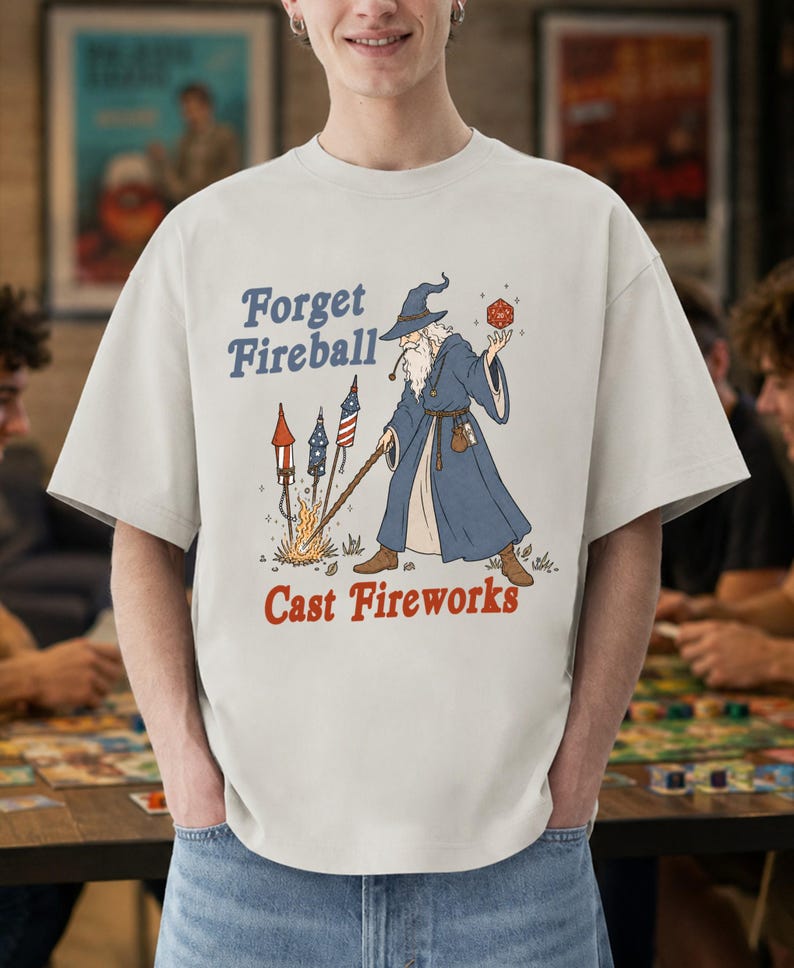 Op de afbeelding: Lichtbeige t-shirt met de tekst "Forget Fireball Cast Fireworks". De afbeelding toont een tovenaar in een blauwe mantel met een rode dobbelsteen en een staf, met vuurwerk. Een casual, unisex stijl.