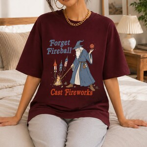 Op de afbeelding: Bordeaux t-shirt met een afbeelding van een tovenaar met een vuurbal en vuurwerk. De tekst op het shirt luidt "Forget Fireball Cast Fireworks" in blauw en oranje. De tovenaar draagt een blauwe jurk en hoed.