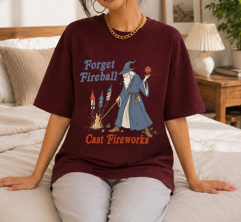 Op de afbeelding: Bordeaux t-shirt met een afbeelding van een tovenaar met een vuurbal en vuurwerk. De tekst op het shirt luidt "Forget Fireball Cast Fireworks" in blauw en oranje. De tovenaar draagt een blauwe jurk en hoed.