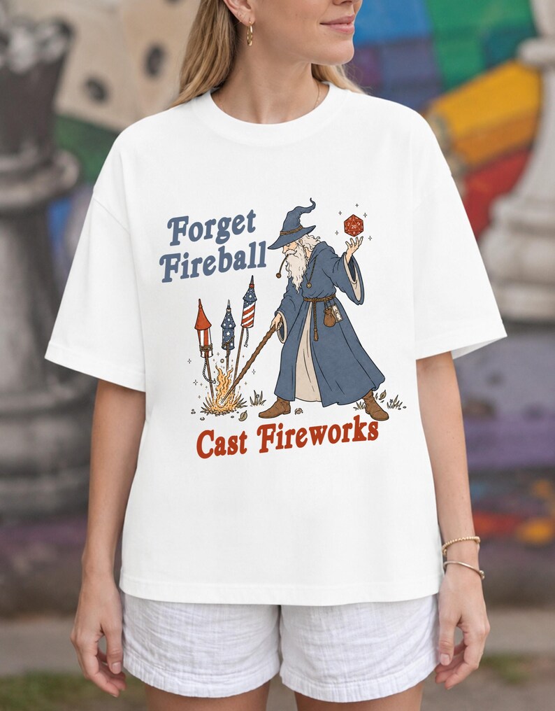 Op de afbeelding: Wit t-shirt met een afbeelding van een tovenaar met een rode dobbelsteen en staf, met vuurwerk en de tekst "Forget Fireball Cast Fireworks" in blauw en rood. De tovenaar draagt een blauwe mantel en hoed.