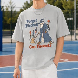 Op de afbeelding: Lichtgrijs t-shirt met een tovenaar afbeelding. De tovenaar houdt een rode bal en een staf vast, met vuurwerk en de tekst "Forget Fireball Cast Fireworks". Het shirt heeft een relaxte, oversized pasvorm.