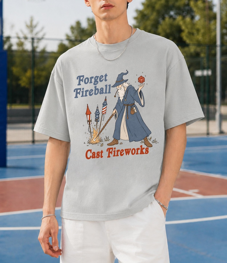Op de afbeelding: Lichtgrijs t-shirt met een tovenaar afbeelding. De tovenaar houdt een rode bal en een staf vast, met vuurwerk en de tekst "Forget Fireball Cast Fireworks". Het shirt heeft een relaxte, oversized pasvorm.