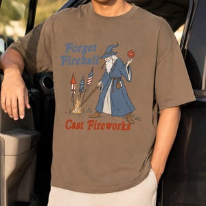 Op de afbeelding: Bruin t-shirt met een afbeelding van een tovenaar met een rode dobbelsteen en een staf, met vuurwerk. De tekst op het shirt luidt "Forget Fireball Cast Fireworks" in blauw en rood. De tovenaar draagt een blauwe jurk en hoed.