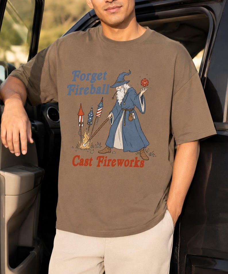 Op de afbeelding: Bruin t-shirt met een afbeelding van een tovenaar met een rode dobbelsteen en een staf, met vuurwerk. De tekst op het shirt luidt "Forget Fireball Cast Fireworks" in blauw en rood. De tovenaar draagt een blauwe jurk en hoed.