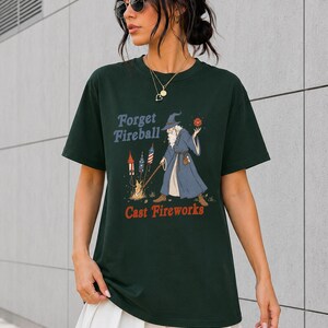 Op de afbeelding: Donkergroen t-shirt met de tekst "Forget Fireball Cast Fireworks" en een tovenaar afbeelding. De tovenaar draagt een blauwe mantel en houdt een rode bol vast. Vuurwerk is afgebeeld aan de linkerkant.