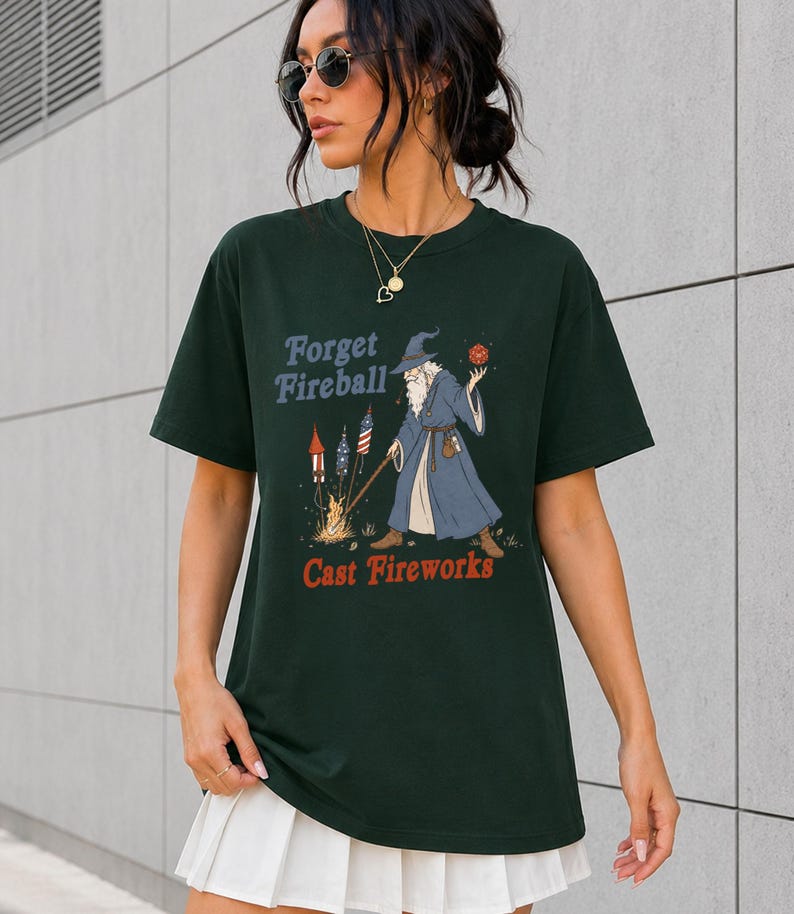 Op de afbeelding: Donkergroen t-shirt met de tekst "Forget Fireball Cast Fireworks" en een tovenaar afbeelding. De tovenaar draagt een blauwe mantel en houdt een rode bol vast. Vuurwerk is afgebeeld aan de linkerkant.
