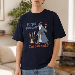 Op de afbeelding: Marineblauw t-shirt met een afbeelding van een tovenaar die vuurwerk afsteekt. De tekst luidt "Forget Fireball, Cast Fireworks" in blauw en rood. De tovenaar draagt een blauwe mantel en houdt een rode dobbelsteen vast.