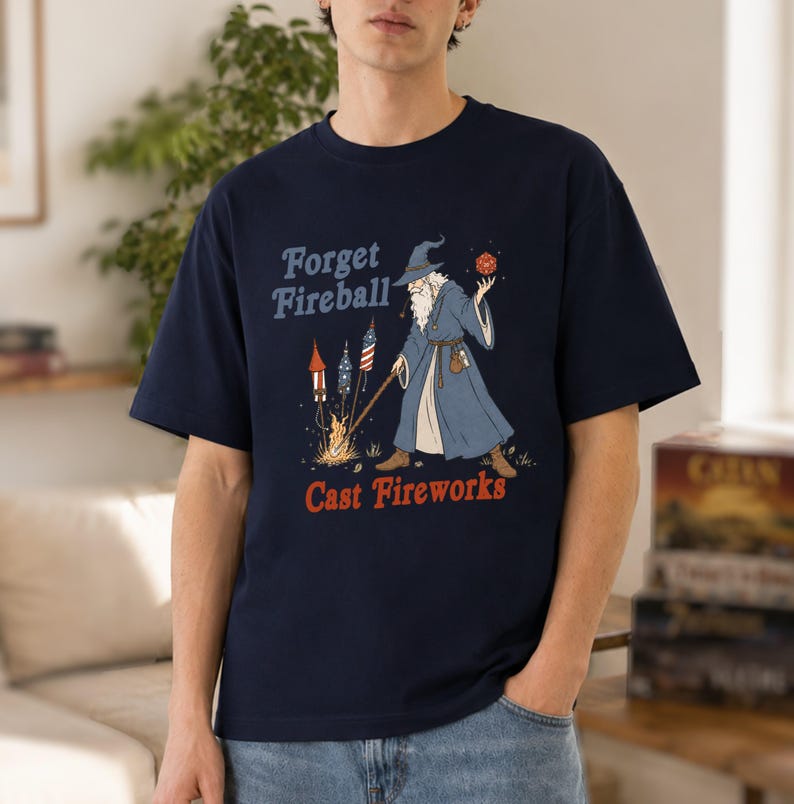 Op de afbeelding: Marineblauw t-shirt met een afbeelding van een tovenaar die vuurwerk afsteekt. De tekst luidt "Forget Fireball, Cast Fireworks" in blauw en rood. De tovenaar draagt een blauwe mantel en houdt een rode dobbelsteen vast.