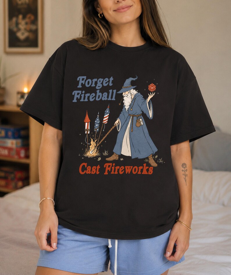 Op de afbeelding: Zwart T-shirt met een afbeelding van een tovenaar met een lange witte baard en een blauwe jurk, met een rode dobbelsteen en een staf. De tekst luidt "Forget Fireball Cast Fireworks" in blauw en rood.