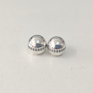 Puede incluir: Dos cuentas esféricas plateadas. Cada cuenta tiene una banda de pequeñas piedras oscuras alrededor del centro. Las cuentas son muy reflectantes, con una superficie lisa y pulida. Joyería.