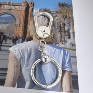 Puede incluir: Un llavero plateado con un mosquet&oacute;n grande y un anillo circular trenzado. El llavero tiene la palabra "PANDORA" grabada en el mosquet&oacute;n y un dise&ntilde;o de corona. El anillo est&aacute; unido al mosquet&oacute;n con una serie de peque&ntilde;os anillos decorativos.