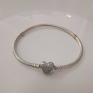 Puede incluir: Una pulsera plateada con un dije en forma de coraz&oacute;n. El dije est&aacute; incrustado con peque&ntilde;as piedras transparentes. La pulsera tiene un dise&ntilde;o de cadena de serpiente y un cierre. Una joya.