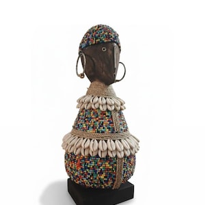 May include: A handcrafted wooden doll with a dark brown face, adorned with a beaded headdress and body. The doll features large hoop earrings and is embellished with seashells and rope detailing. It stands on a black square base.