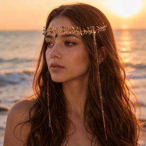 May include: A gold-toned metal headpiece featuring a delicate leaf-patterned band adorned with small green accents. A central teardrop-shaped green stone hangs from the center, while two long, thin gold chains drape down from the sides of the band.