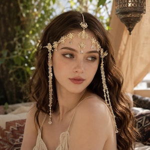May include: A gold-toned headpiece featuring intricate filigree metalwork and dangling clear crystal beads. The accessory rests across the forehead with a central chain extending upward and long beaded strands draping down along the sides.