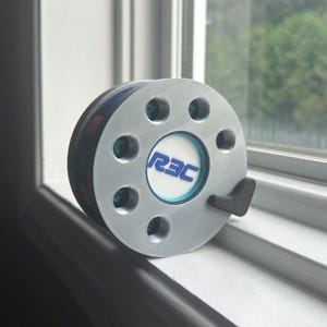 Peut inclure: Un dispositif circulaire de couleur argentée avec un disque central blanc portant le texte bleu R3C. L'objet présente huit découpes circulaires régulièrement espacées autour du centre et un petit levier noir en forme de L sur le bord extérieur.