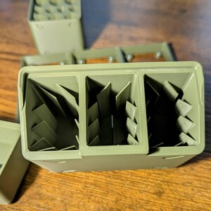 May include: Olive green 3D-printed organizer with three compartments, each featuring angled dividers. The organizer is rectangular with a textured surface. Additional matching organizers are visible in the background, suggesting a modular design for storage and organization.