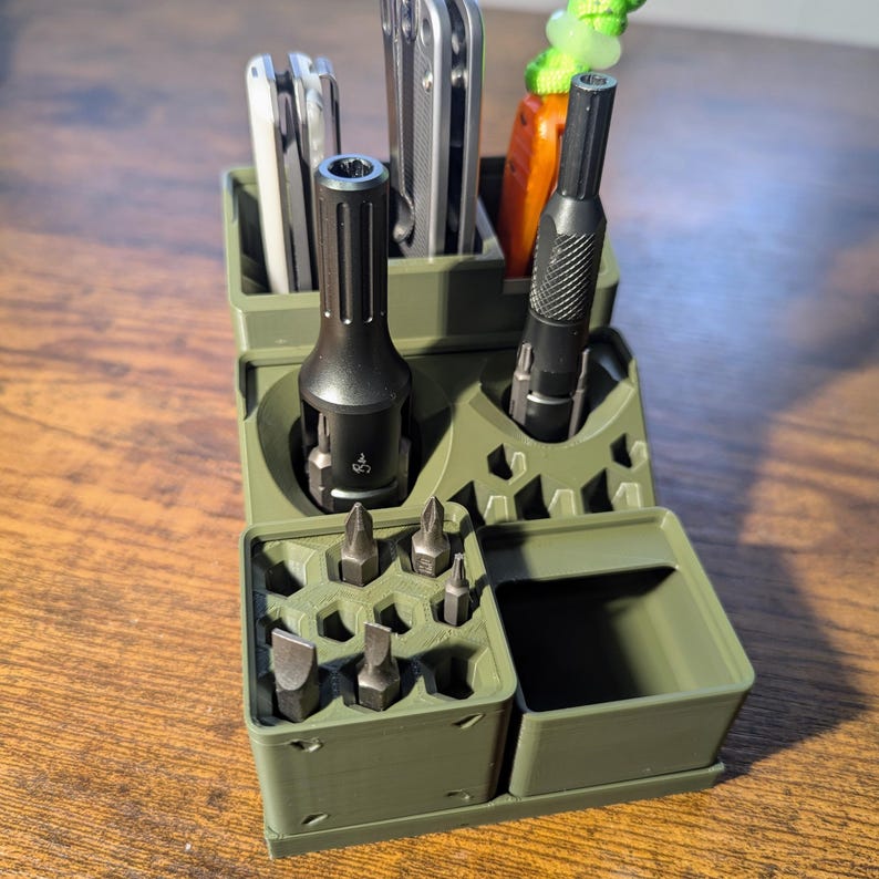 May include: An olive green organizer with multiple compartments holding tools. The organizer has slots for screwdrivers, bits, and other tools. A black screwdriver and a multi-tool are visible. The organizer is made of plastic and sits on a wooden surface.