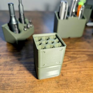 May include: A set of olive green 3D-printed organizers. The primary organizer is a square container with a honeycomb top, designed to hold tools. Other organizers hold various tools, including screwdrivers and pens. The items are arranged on a wooden surface.