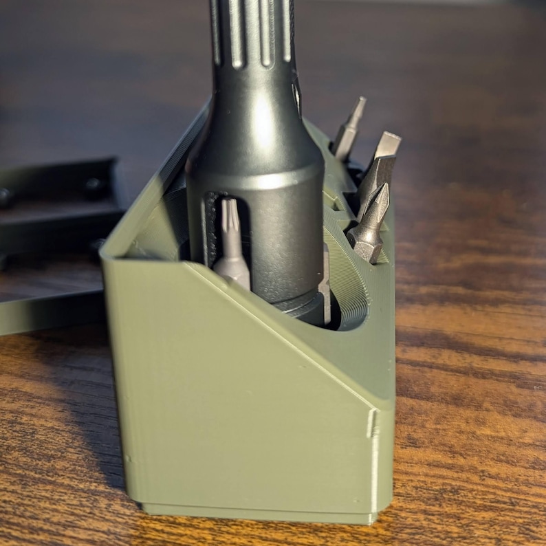 May include: An olive green tool organizer holding a dark gray screwdriver and several silver bits. The organizer has a rectangular shape with angled sides and slots for the bits. The screwdriver has a textured handle.