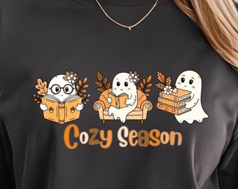 Könnte beinhalten: Schwarzes T-Shirt mit einer Grafik von drei Geistern, die Bücher lesen, mit dem Text "Cozy Season" in Orange. Die Geister sind mit Blättern und Brillen dargestellt, sitzen auf einem Stuhl und halten Bücher. Das Shirt ist ein lässiger, übergroßer Stil.