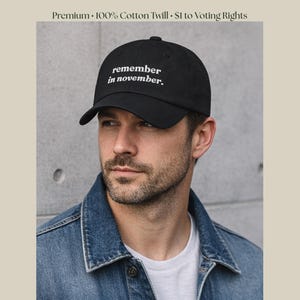 Könnte beinhalten: Schwarze Baseballkappe mit weißem Schriftzug "remember in november". Die Kappe besteht aus 100 % Baumwoll-Twill. Die Person im Bild trägt eine Jeansjacke und ein weißes T-Shirt.