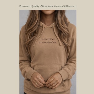 May include: A tan hoodie with a front pocket and drawstring hood. The text "remember in november" is embroidered in a darker brown color on the chest. The hoodie is made of a soft material.