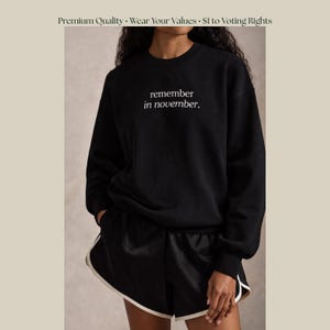 May include: Black sweatshirt with the text "remember in november" in white. The model is also wearing black shorts with white trim. The image has the text "Premium Quality Wear Your Values. $1 to Voting Rights" at the top.