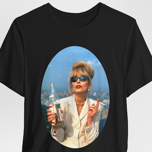 Puede incluir: Camiseta negra con una imagen impresa de una mujer con gafas de sol sosteniendo una botella y un cigarrillo. La imagen está enmarcada en un óvalo contra un fondo de paisaje urbano. La mujer lleva un traje blanco y joyas de oro.