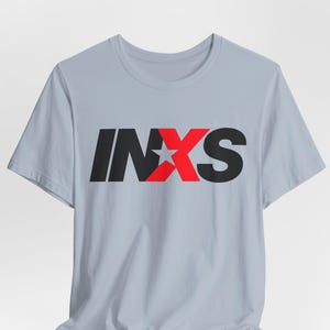 May include: A light gray short-sleeve t-shirt featuring the text INXS printed across the chest. The letters I, N, and S are black, while the letter X is red and contains a white five-pointed star in the center of the character.