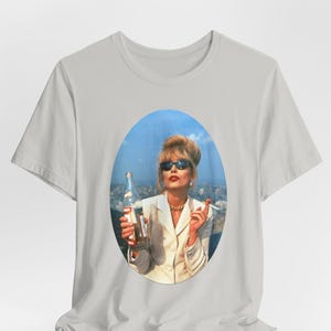 Może przedstawiać: Jasnoszary t-shirt z krótkim rękawem z centralnym owalnym nadrukiem graficznym przedstawiającym osobę o blond włosach w ciemnych okularach przeciwsłonecznych i białej marynarce, trzymającą szklaną butelkę i papierosa na tle miasta.