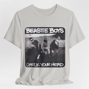 Puede incluir: Camiseta gris claro con una imagen en blanco y negro de los Beastie Boys. La imagen incluye el texto "BEASTIE BOYS" arriba y "CHECK YOUR HEAD" abajo.