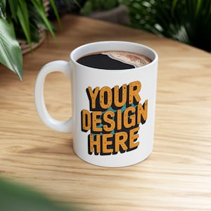 Puede incluir: Taza de cerámica blanca llena de café oscuro, con el texto "YOUR DESIGN HERE" en letras de bloque naranja y negro. La taza tiene un asa blanca y está sobre una superficie de madera clara.