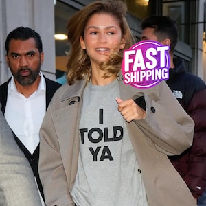 Puede incluir: Una camiseta gris claro con el texto "I TOLD YA" en letras negras. La persona lleva una gabardina color beige. Es visible una pegatina morada y rosa que dice "FAST SHIPPING".