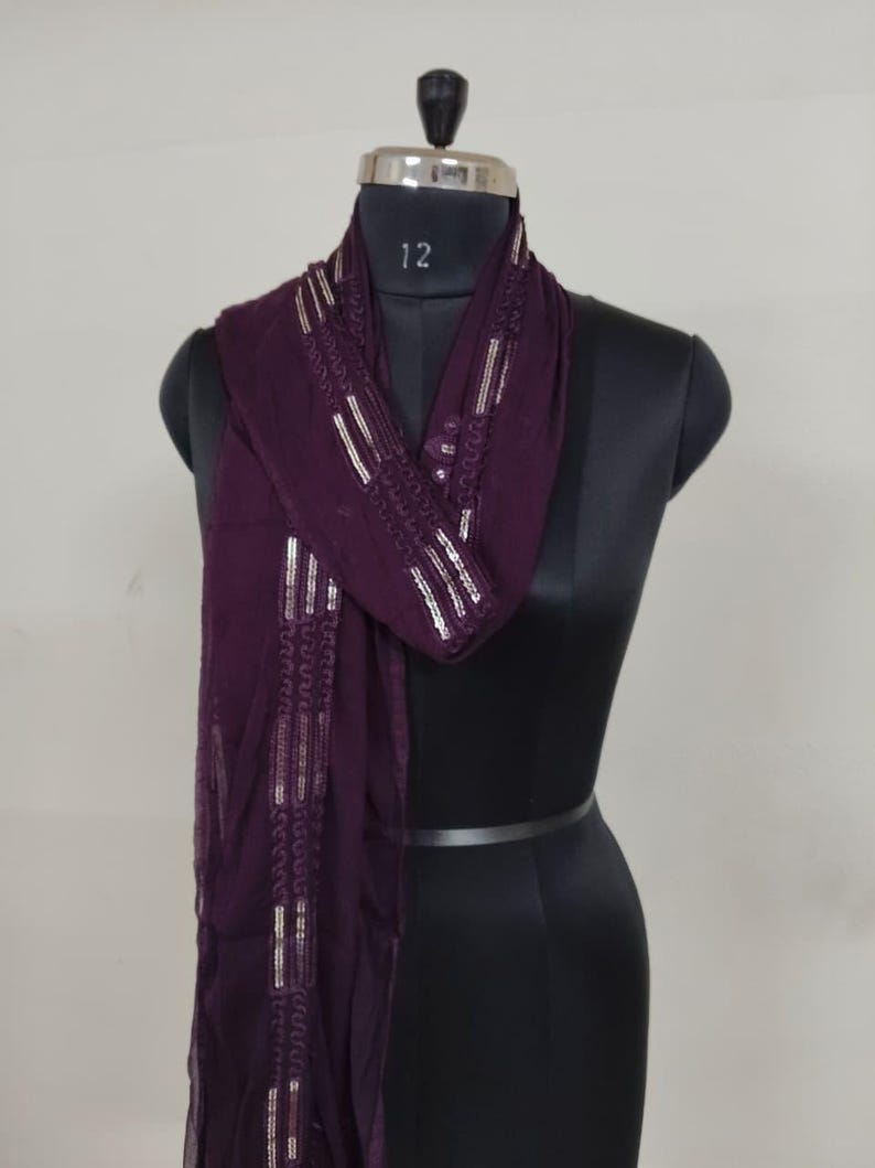 May include: A dark purple scarf with silver embellishments is draped over a black mannequin. The scarf has a rectangular shape with decorative silver lines and patterns along the edges. The mannequin has a silver neck piece with the number 12.