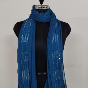 May include: A teal scarf with intricate sequin and embroidered detailing. The scarf is draped over a black mannequin, showcasing its flowing design. The fabric appears lightweight and suitable for layering.