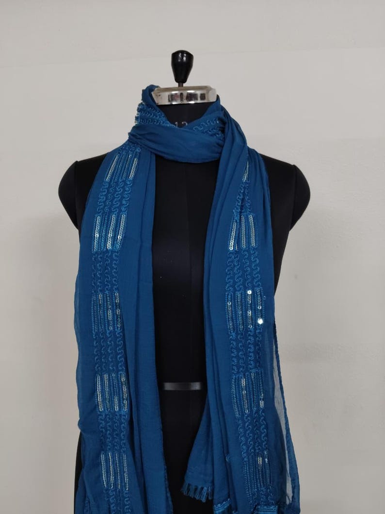 May include: A teal scarf with intricate sequin and embroidered detailing. The scarf is draped over a black mannequin, showcasing its flowing design. The fabric appears lightweight and suitable for layering.