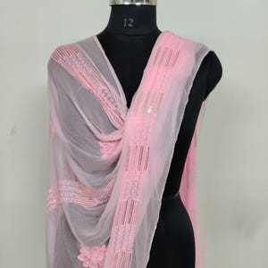 May include: A sheer, light pink scarf with intricate embroidered details and sequin accents. The scarf is draped over a black mannequin, showcasing its delicate floral and geometric patterns. The fabric appears lightweight and flowing.